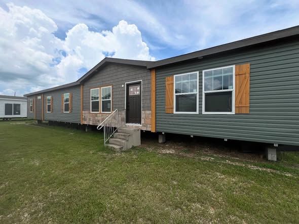 Modern ranch-style modular home exterior featuring dark sage green horizontal siding, decorative stone accents around the entryway, and rustic wooden shutters on large white-trimmed windows. The architectural design includes a gabled roofline with grey shingle accents and a simple concrete staircase leading to a dark brown front door, all situated on a grassy lot under a bright, cloudy sky to showcase a contemporary prefab housing aesthetic.