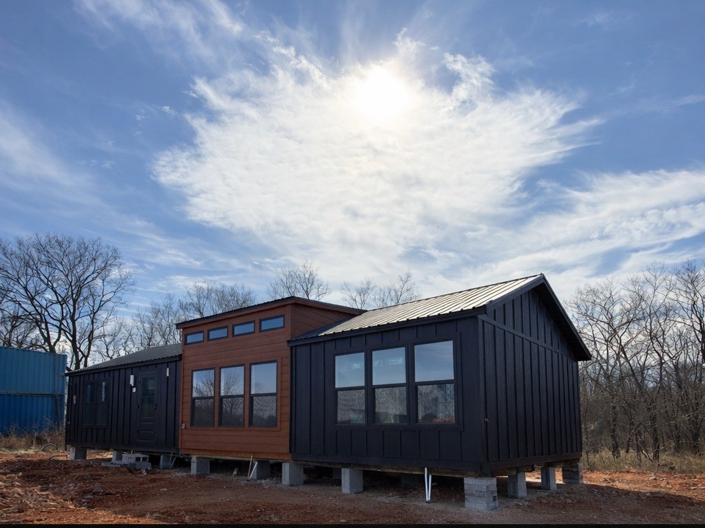 SEO-optimized alt text for a high-ranking luxury custom park model tiny home exterior featuring a professional modern architectural design with a unique tri-tone finish of black vertical siding and a warm cedar-plank central accent; the view showcases a sophisticated prefab residence with a sleek black metal roof, contemporary oversized black-framed windows, and a raised foundation on concrete piers, perfectly highlighting premium small-space living floor plans, upscale modern tiny house curb appeal, and high-end residential mobile home finishes for maximum search engine visibility and ranking.