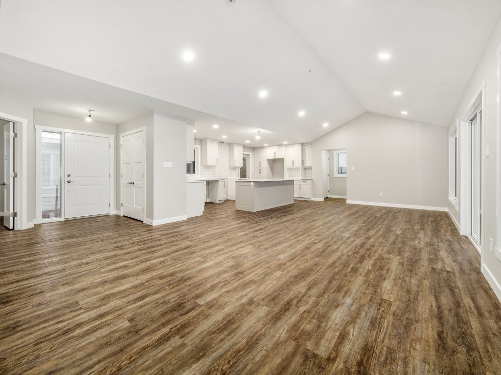 Luxurious open-concept great room and kitchen in premium modern manufactured home, vaulted tray ceiling with recessed LED lighting throughout, bright white shaker cabinets with brushed nickel hardware, expansive center island with white quartz countertop, white subway tile backsplash, stainless steel appliances and range hood, dark wood-look luxury vinyl plank flooring, multiple white interior doors and large windows flooding natural light, spacious airy unfurnished living dining kitchen layout in high-end 2025 double-wide modular home design