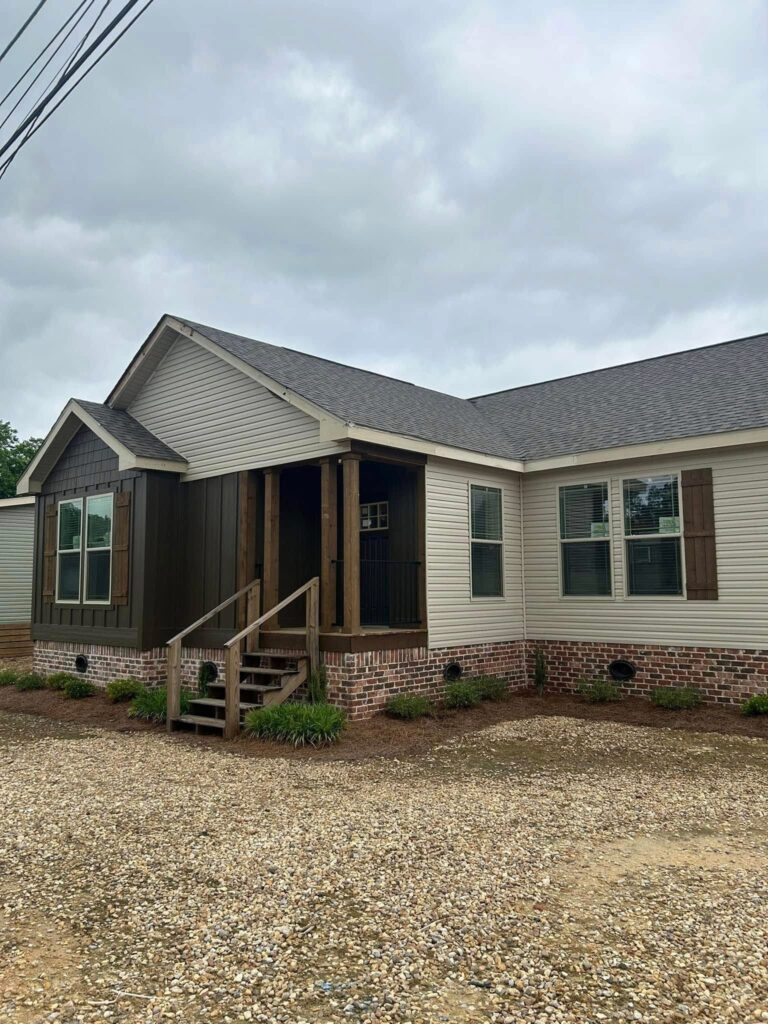 Exterior view of a modern manufactured home featuring a welcoming covered porch with wooden beams, a dual-tone siding exterior in beige and dark brown, a brick foundation, and wooden shutters on the windows, set against a backdrop of a cloudy sky and a gravel driveway.