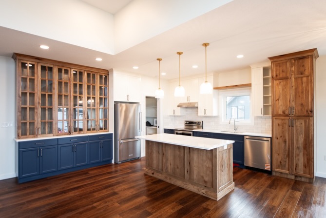 SEO-optimized alt text for a high-ranking luxury custom modular home kitchen featuring a professional open-concept interior design with a striking navy blue and rustic wood cabinetry combination; the architectural view showcases a large wood-plank kitchen island with a white quartz countertop, a grand floor-to-ceiling glass-front hutch, and premium stainless steel appliances, all complemented by elegant white shaker upper cabinets, a classic subway tile backsplash, and contemporary gold pendant lighting set against rich dark wide-plank hardwood flooring, perfectly highlighting gourmet prefab kitchen layouts, modern craftsman-style architectural details, and high-end residential mobile living solutions for maximum search engine visibility and ranking.