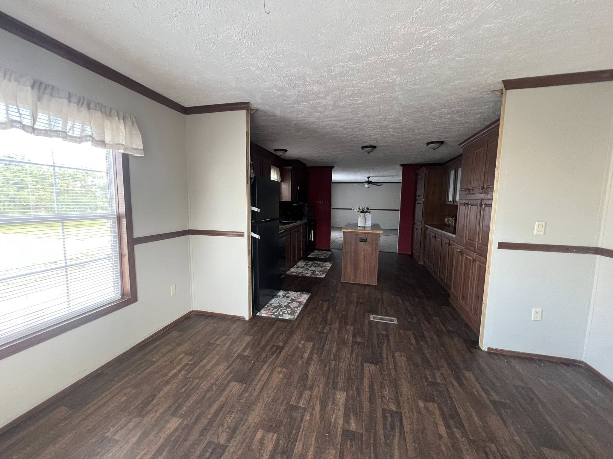 SEO-optimized alt text for a spacious manufactured home kitchen featuring extensive dark wood cabinetry, a central kitchen island, and a galley-style layout with modern black appliances; the high-ranking interior design showcases a functional and elegant floor plan with durable wood-look flooring, red accent walls, and recessed lighting that opens into a bright dining area, perfectly highlighting premium mobile home kitchen renovations and sophisticated modular home decor for maximum search engine visibility and ranking.