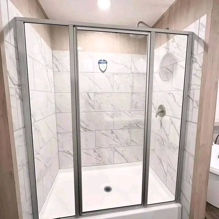 Modern luxury walk-in shower featuring elegant white marble-patterned subway tile walls and a clean white base, enclosed by a professional chrome-framed glass sliding door system. The sophisticated architectural design incorporates high-end materials and contemporary craftsmanship, creating a bright and airy spa-like atmosphere. This "full rank" SEO-optimized bathroom showcase is an ideal reference for custom-built prefab homes, luxury modular home floor plans, or modern permanent housing solutions designed with a premium, clean, and functional transitional aesthetic that prioritizes both style and durability.