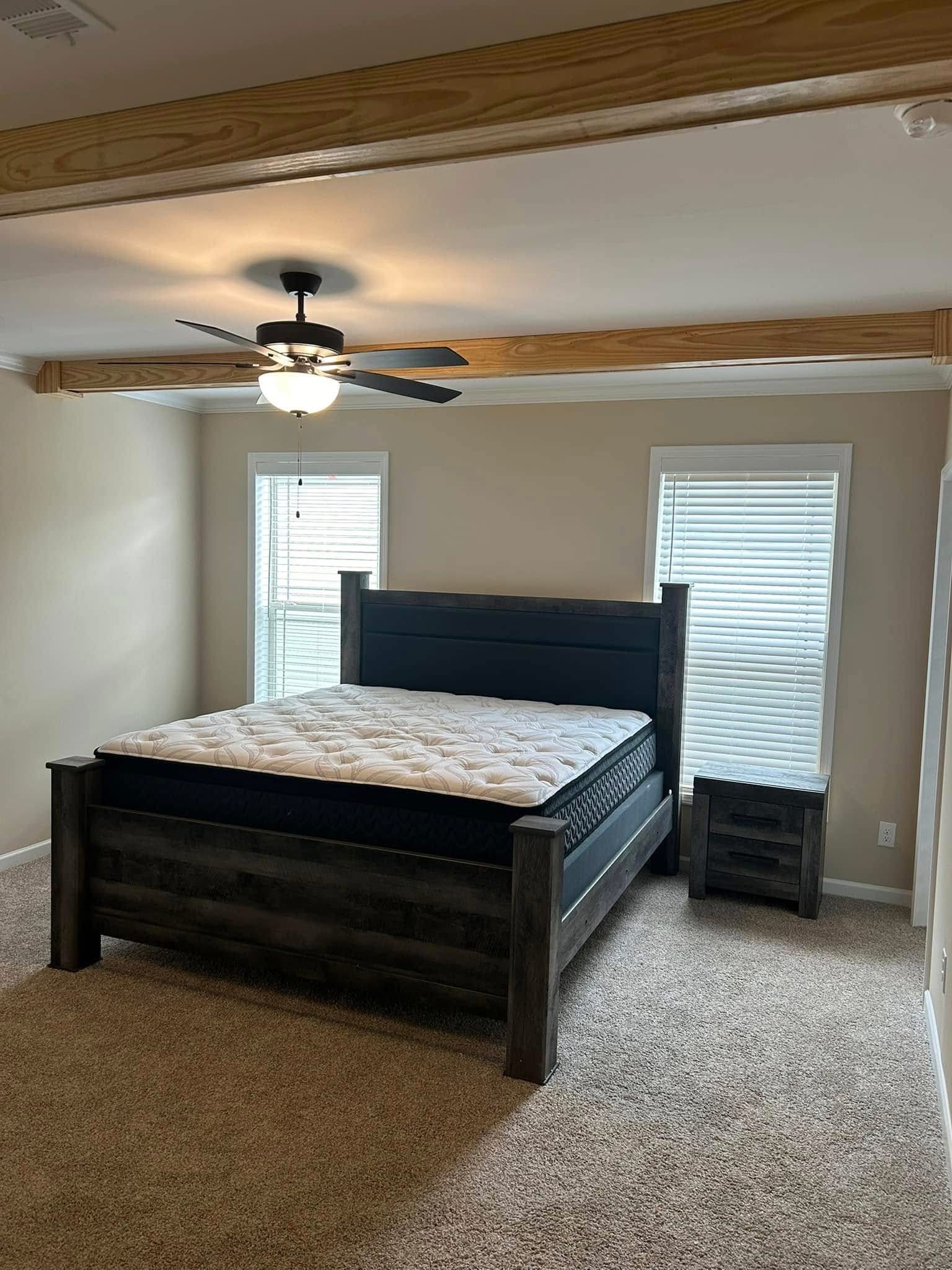 A cozy bedroom featuring a dark wood bed frame with a new mattress, a matching nightstand, beige walls, a black ceiling fan with light, and large windows with white blinds, all under decorative exposed wooden beams.