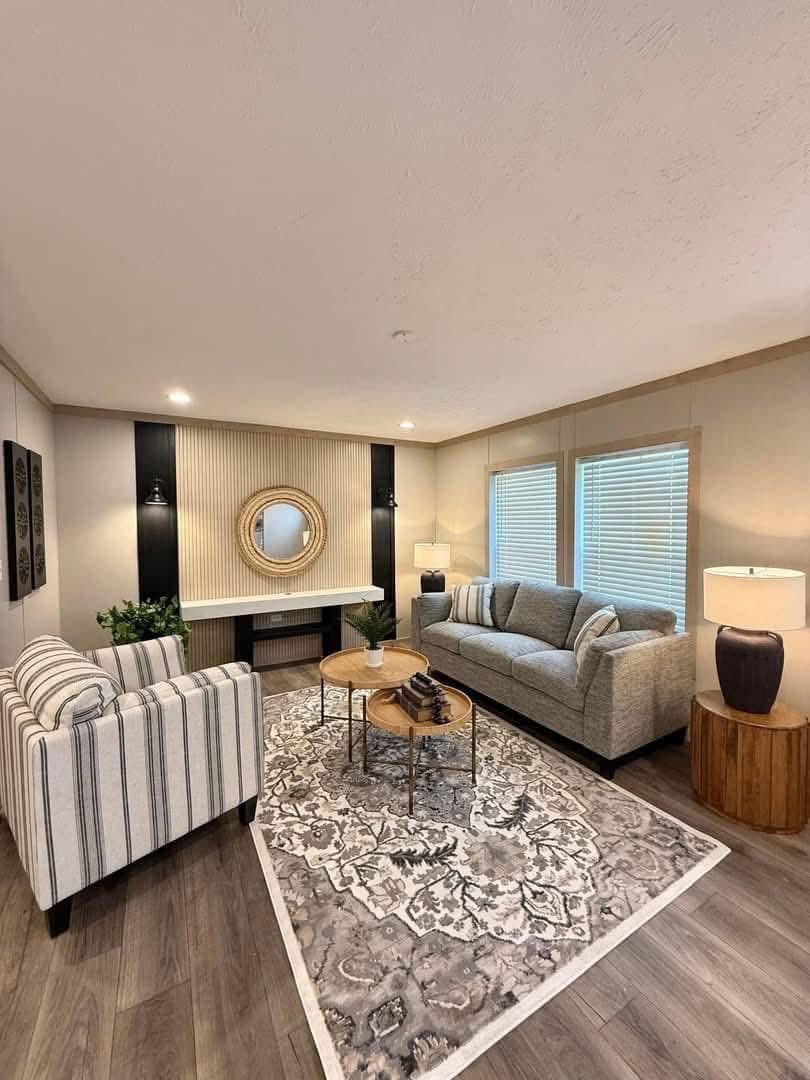 Modern luxury manufactured home interior featuring a stylish transitional living room with a grey sofa and striped armchair, centered around a vintage floral area rug and nested wooden coffee tables, complemented by a unique slatted accent wall with a circular mirror and integrated lighting, all set on high-end wood-look vinyl flooring for a sophisticated move-in ready residential design.