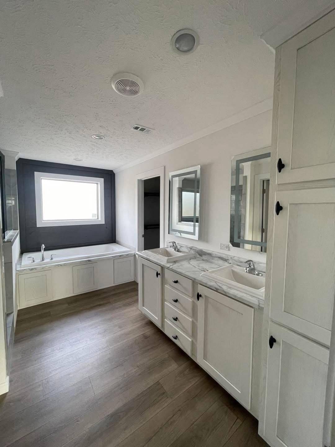 Spacious bathroom with wood floors, white double-sink vanity with marble top, large tub under a window, and light gray walls creating a calm, clean atmosphere.