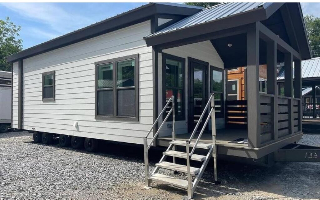 Modern luxury single-wide manufactured home exterior featuring a sleek white horizontal siding design with dark charcoal trim, a spacious covered front porch with a gabled roof and integrated privacy slats, and a durable multi-axle mobile chassis, showcasing a high-end move-in ready turn-key residential property for a sophisticated tiny home lifestyle.