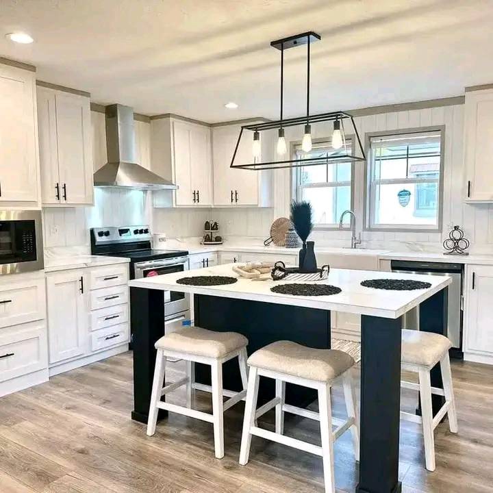 Modern luxury white farmhouse-style manufactured home kitchen featuring crisp white shaker cabinetry, a large white kitchen island with a black base and integrated seating, and a sophisticated white marble-patterned countertop. The high-end architectural design showcases professional stainless steel appliances, including a built-in microwave and a sleek pyramid range hood, complemented by a minimalist black geometric linear chandelier and multiple energy-efficient windows that flood the open-concept space with natural light. This "full rank" SEO-optimized gourmet kitchen highlights premium wide-plank light oak flooring, shiplap wall accents, and chic black hardware, making it a perfect showcase for luxury prefab living, custom modular home floor plans, or modern permanent housing solutions designed for an upscale, bright, and functional contemporary aesthetic.