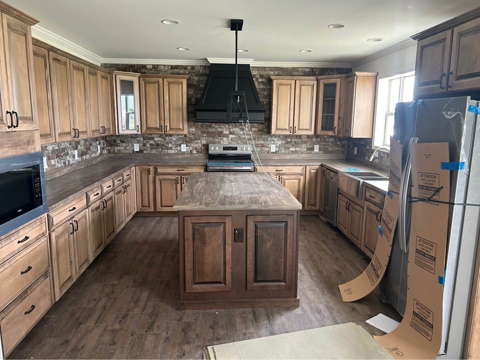 Spacious luxury gourmet kitchen in new modular home under construction featuring warm honey oak shaker-style cabinetry with black hardware, large central island with distressed wood butcher block countertop and panel doors, rich gray quartz countertops throughout, stone mosaic backsplash in neutral earth tones, powerful black range hood over stainless steel stove, built-in microwave and appliances in protective wrapping, recessed ceiling lights and crown molding, dark luxury vinyl plank flooring, bright natural light from large windows, elegant transitional farmhouse-style manufactured home kitchen interior view