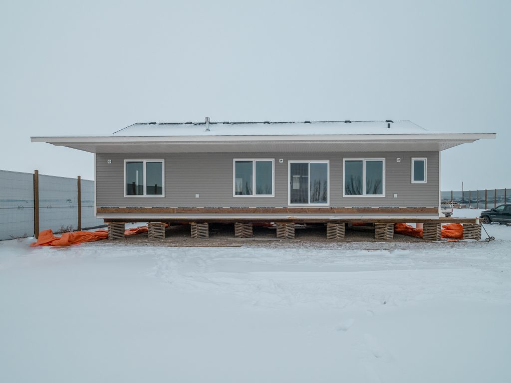 Modern single-story manufactured home exterior in winter snowy landscape, sleek gray vertical siding with white trim and wide overhanging flat roof eaves, large energy-efficient windows including sliding glass patio doors, white-framed glass entry door, elevated on temporary wooden foundation beams and concrete blocks with orange protective tarps and construction materials around, newly delivered modular home ready for setup in cold climate rural setting, contemporary energy-efficient double-wide mobile home 2025 model under overcast sky
