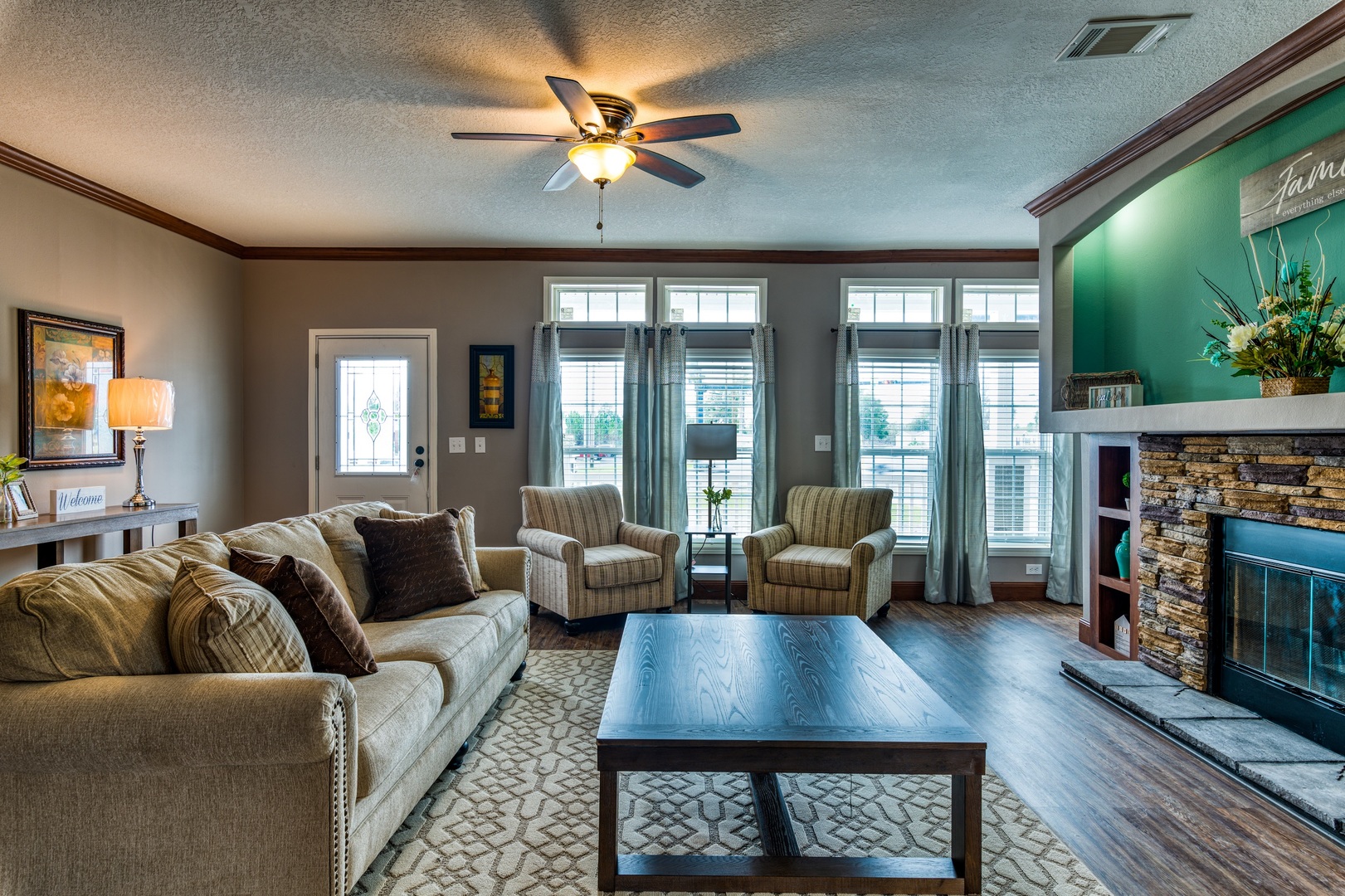 A comfortable living room with a beige sectional sofa, two striped armchairs, a dark wood coffee table, a stone fireplace with a teal accent wall, and large windows with blue curtains.