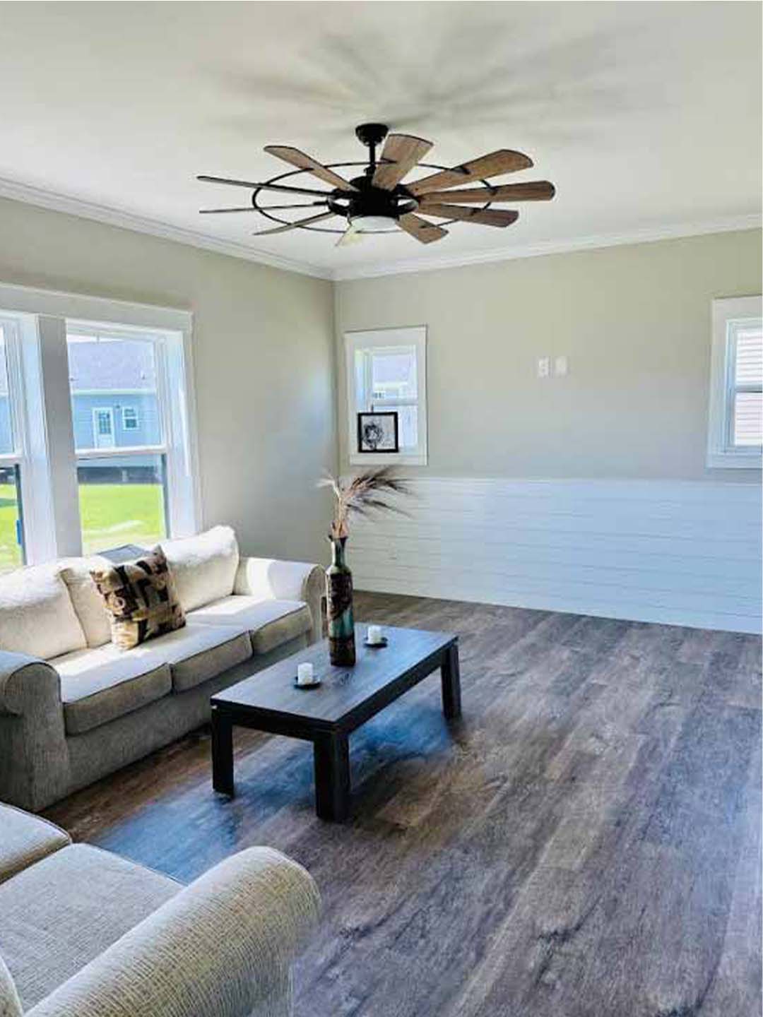 Modern luxury kitchen and dining room interior featuring bright white shaker cabinets with black hardware, high-end stainless steel appliances, and a large white marble-topped kitchen island with industrial black pendant lighting. The open-concept design is enhanced by grey wide-plank wood-look flooring, a white subway tile backsplash, and a large window providing natural light, while a dark wood ceiling beam adds a rustic touch to this sophisticated contemporary farmhouse-style home.