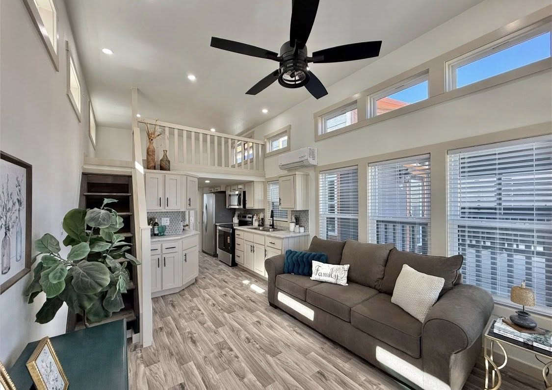 Bright, modern tiny home interior with high ceiling, ceiling fan, and loft. Features a kitchen, L-shaped cabinets, sofa, large windows, and neutral decor.