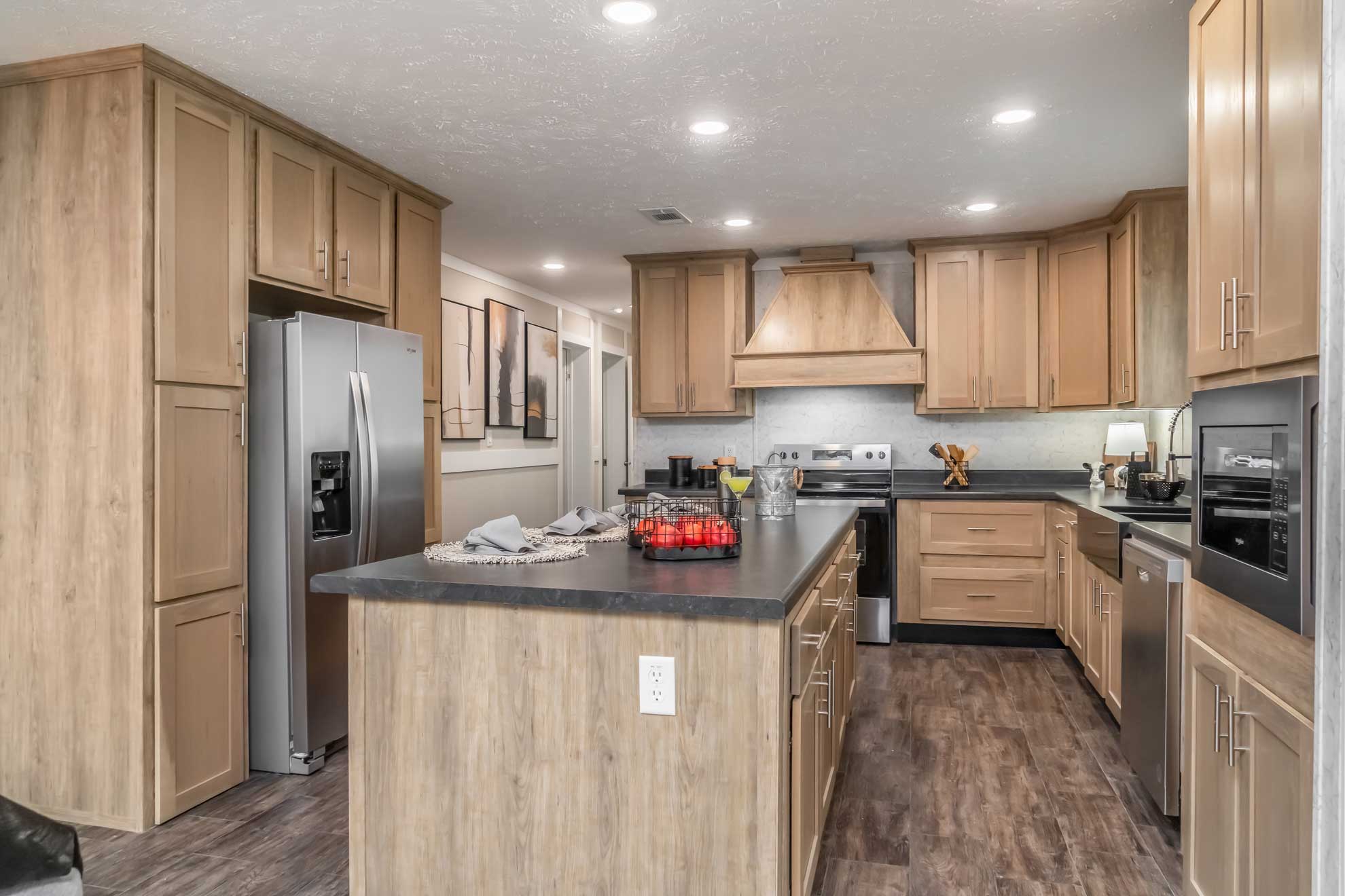 SEO-optimized alt text for a modern, large manufactured home kitchen featuring light oak-finished cabinetry, a large central island with a black slate-style countertop, and premium stainless steel appliances; the high-ranking interior design showcases a functional open-concept layout with a matching wood vent hood, marble-patterned backsplash, and durable wood-plank flooring, perfectly highlighting luxury modular home kitchen renovations and contemporary mobile home floor plans for maximum search engine visibility and ranking.
