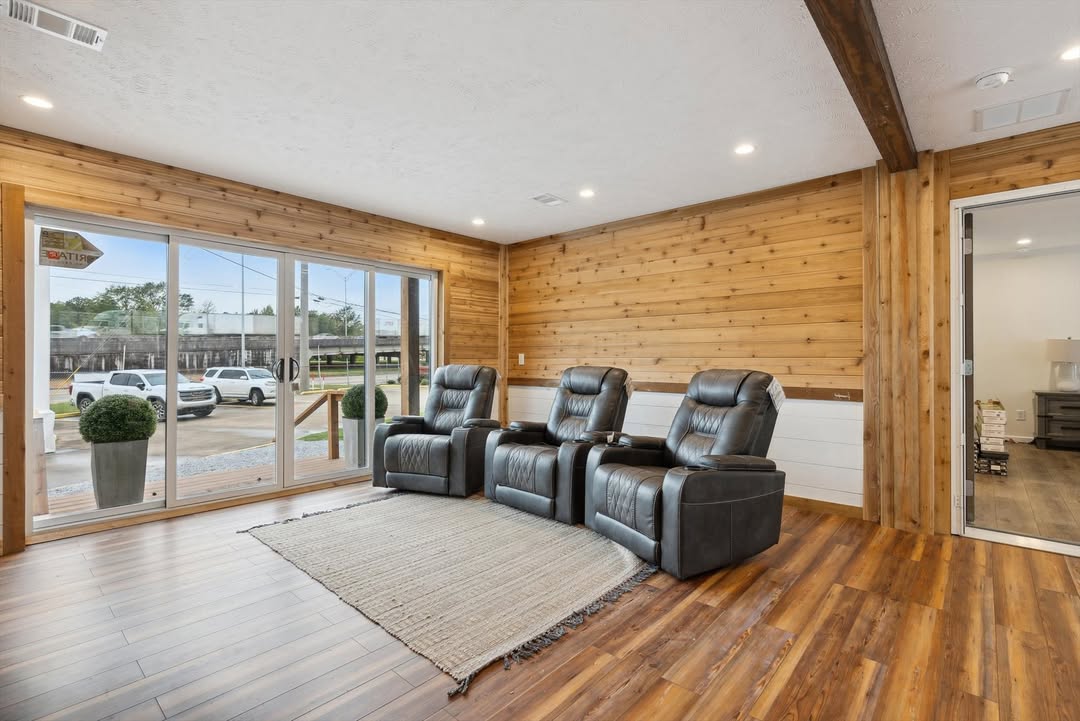 Cozy modern home theater lounge in premium modular manufactured home featuring three plush black leather power recliners arranged in front of large floor-to-ceiling sliding glass patio doors with scenic outdoor views, rustic horizontal wooden plank accent walls in warm honey tones, exposed dark wooden ceiling beams with recessed lighting, textured vaulted ceiling, light gray area rug over wide-plank luxury vinyl flooring, bright airy sunroom-style media room interior with natural daylight flooding in, elegant transitional farmhouse design perfect for family movie nights