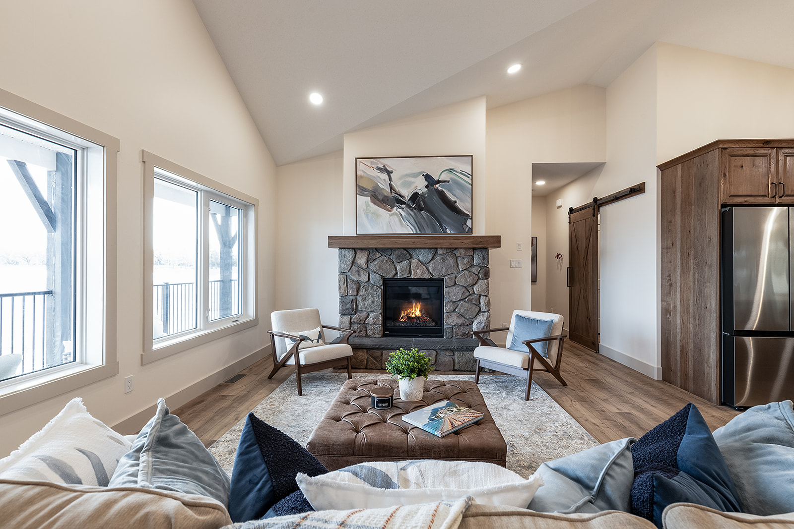 Cozy living room with a stone fireplace, abstract art, and armchairs. A plush ottoman and cushions enhance warmth. Natural light streams in through large windows.
