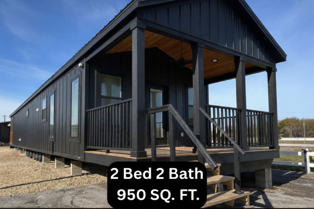 A modern, black tiny house with a gable roof and front porch sits on gravel. Text reads "2 Bed 2 Bath, 950 SQ. FT." Clear blue sky in the background.