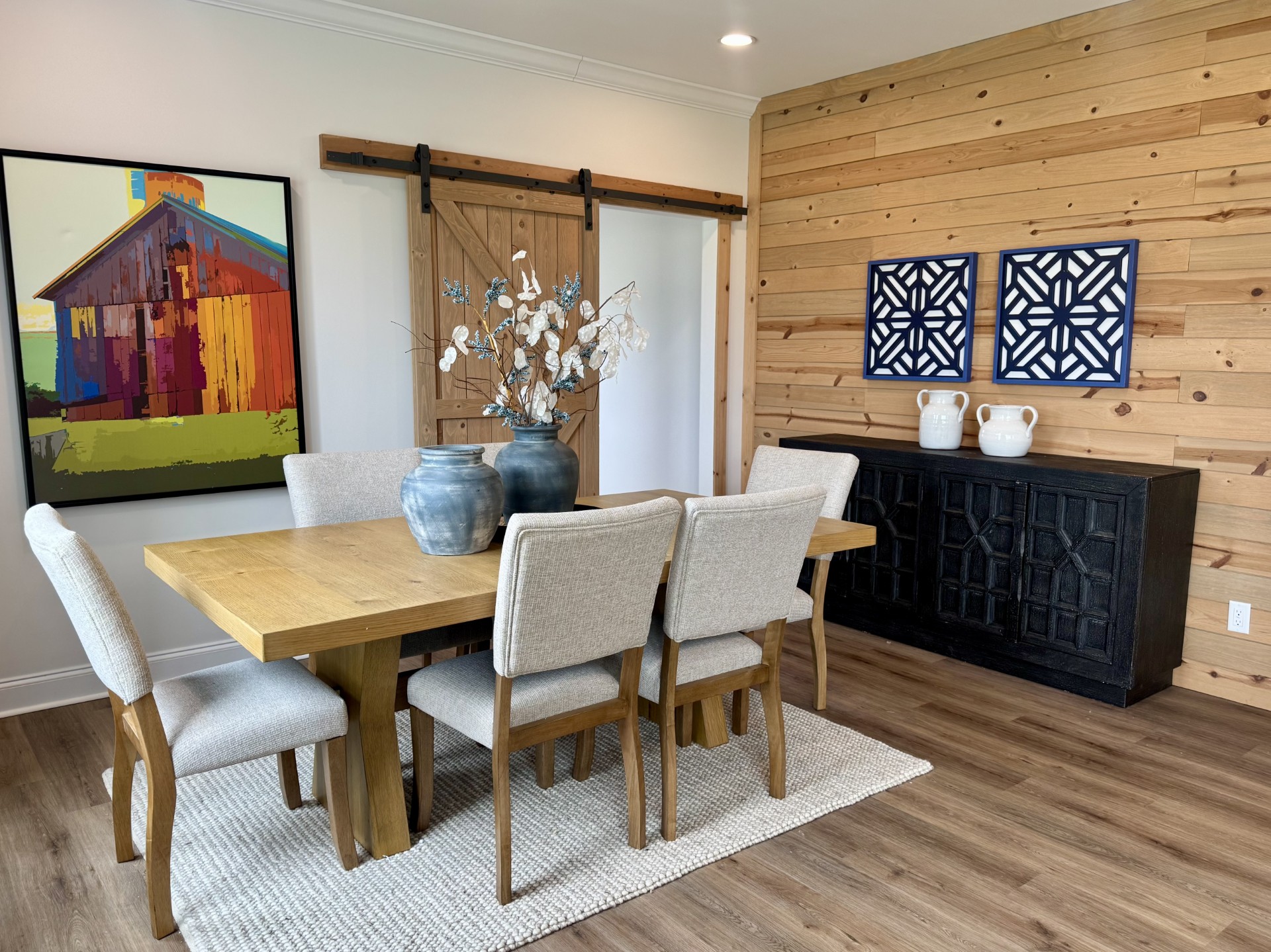 SEO-optimized alt text for a high-ranking luxury custom modular home dining room featuring a professional rustic-modern interior design with a warm wood-plank accent wall and a sliding barn door; the architectural view showcases a solid light-oak dining table with neutral upholstered chairs set on a woven cream area rug, complemented by a large vibrant barn-style art piece, a sophisticated black geometric sideboard, and decorative blue ceramic vases, all set against durable wide-plank flooring to highlight luxury prefab home floor plans, modern farmhouse-style dining decor, and high-end residential mobile living solutions for maximum search engine visibility and ranking. Would you like me to create the alt text for the living room or the blue kitchen next?