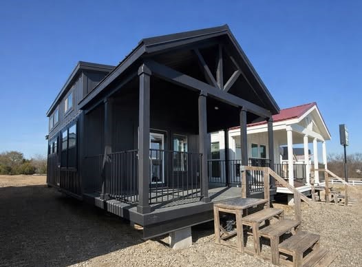 A modern tiny house with a dark exterior and covered porch, standing on gravel with clear blue skies. Steps lead up to the entrance, evoking a cozy and inviting feel.