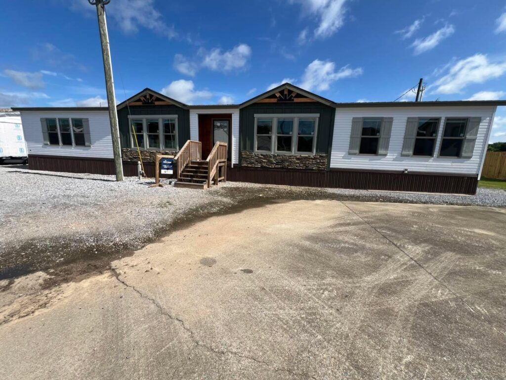 Modern luxury double-wide manufactured home for sale featuring a triple-gabled front exterior with white horizontal siding, forest green accent panels, and rustic stone skirting, complete with a natural wood entry staircase and expansive multi-pane windows for a sophisticated, high-end, and move-in ready turn-key residential property.