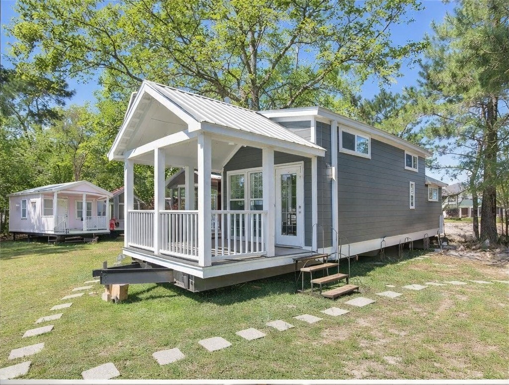 SEO-optimized alt text for a high-ranking luxury park model tiny home exterior featuring a professional architectural design with a gabled metal roof and a spacious vaulted front porch; the view showcases a modern dark gray prefab residence with crisp white trim, a contemporary white railing, and a welcoming glass-paneled front door, complemented by a rustic stone-step walkway and a lush wooded background to highlight premium small-space living floor plans, upscale tiny house curb appeal, and luxury residential mobile home finishes for maximum search engine visibility and ranking.