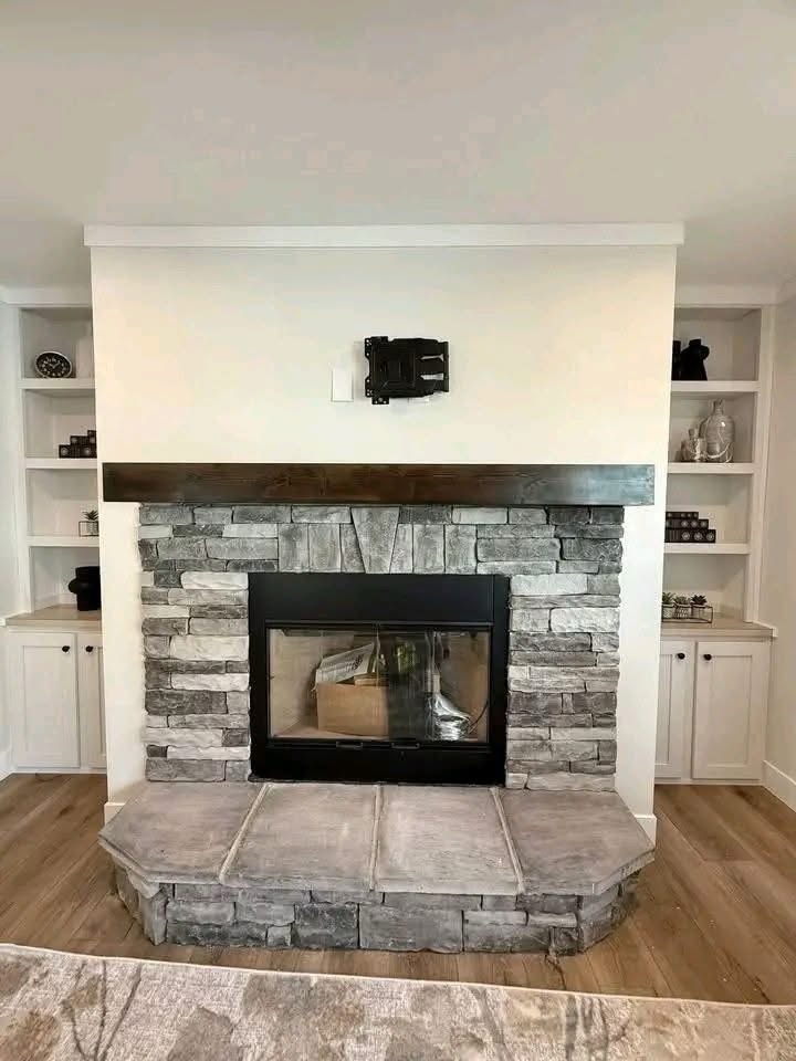 
Gemini said
Modern living room featuring a floor-to-ceiling stone fireplace with a dark wood mantle, integrated black electric firebox, and a raised grey stone hearth, flanked by custom white built-in bookshelves and cabinets on light oak hardwood flooring, perfect for contemporary home interior design and cozy rustic living space inspiration.