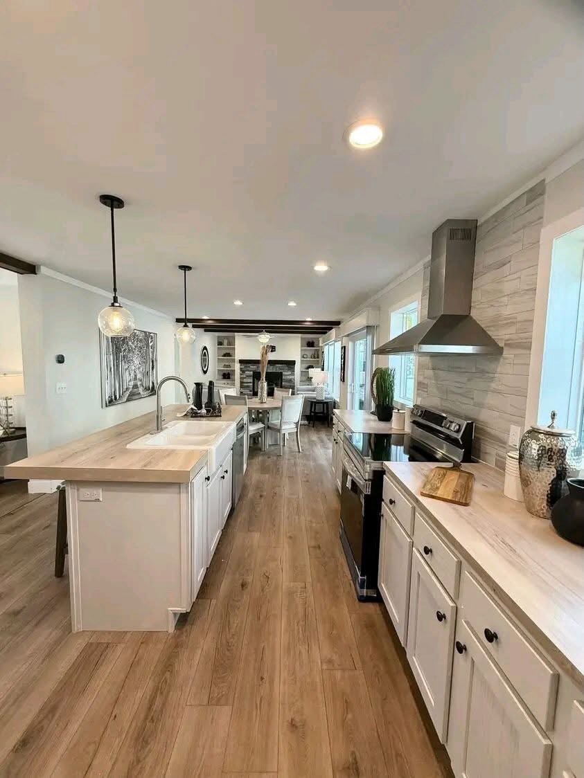 
Gemini said
Modern open-concept galley kitchen featuring white shaker cabinets, light oak wood countertops, and a large kitchen island with a white porcelain farmhouse sink, equipped with stainless steel appliances and industrial pendant lighting, flowing into a bright dining area and living room with dark wood ceiling beams and a stone fireplace for a cohesive transitional home interior design.