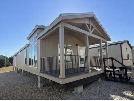 Modern luxury prefab tiny home with elevated foundation on concrete piers, spacious wraparound porch featuring black metal railings and sturdy columns, vaulted gable roof with exposed beams, large panoramic floor-to-ceiling windows for maximum natural light, light taupe vertical board and batten siding, contemporary farmhouse exterior design, spacious deck overlooking scenic landscape, ideal for off-grid living, modular tiny house on wheels alternative, high-end manufactured home with premium finishes and countryside views