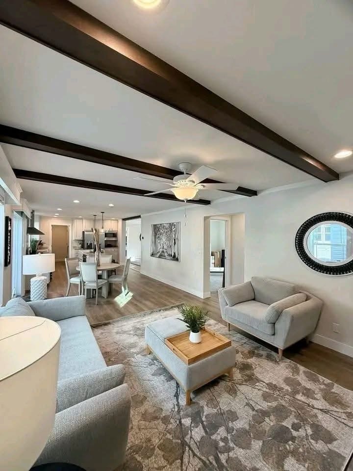 
Gemini said
Modern open-concept living area and kitchen featuring dark wood ceiling beams, a light grey sectional sofa and matching armchair on a floral area rug, and a white farmhouse sink on a butcher block island, creating a cohesive and bright transitional home interior design perfect for modern family living.