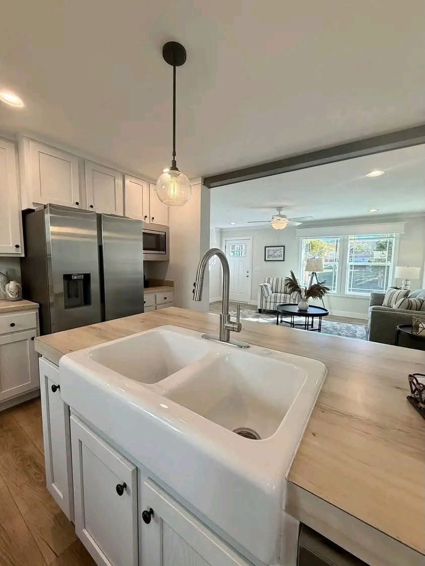 
Gemini said
Modern open-concept kitchen featuring a white farmhouse drop-in sink with a brushed nickel gooseneck faucet on light wood countertops, stainless steel appliances, and white cabinetry, providing a clear view into a bright living room with neutral decor, a striped armchair, and large windows for a perfect home renovation and interior design inspiration.