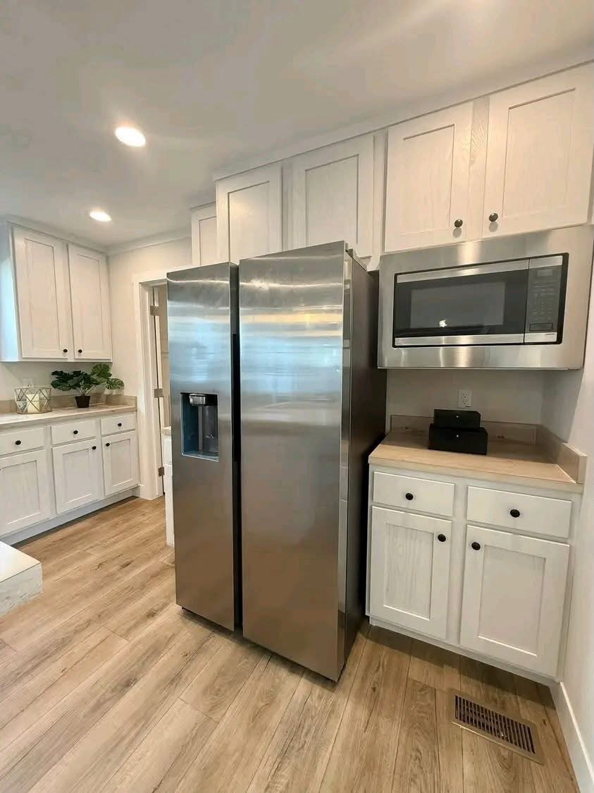 
Gemini said
Modern open-concept galley kitchen and living room featuring white shaker cabinets, light oak butcher block countertops, a white double-bowl farmhouse sink with a brushed nickel gooseneck faucet, and stainless steel appliances, seamlessly transitioning into a bright living area with dark wood ceiling beams, neutral grey furniture, and a stone fireplace for a cohesive transitional home interior design.