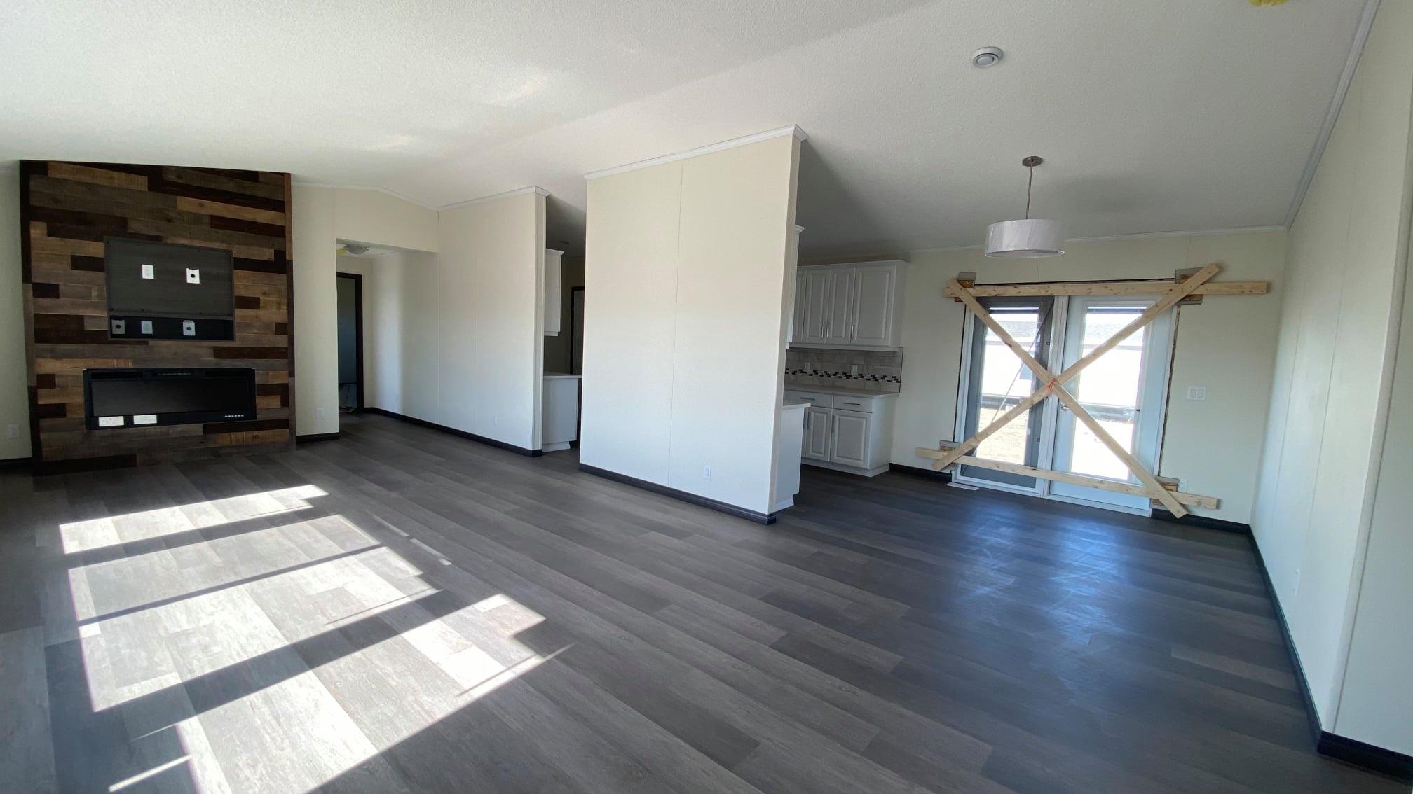 Spacious open-concept great room and kitchen interior of a new modular home under construction with large sliding glass doors, natural sunlight pouring in, feature accent wall with multi-toned reclaimed wood paneling surrounding modern electric fireplace and wall-mounted TV, light gray luxury vinyl plank flooring throughout, vaulted textured ceiling, white shaker kitchen cabinets with island, pendant light fixtures, neutral beige walls, temporary wooden bracing on patio doors, empty unfurnished modern manufactured home living space view