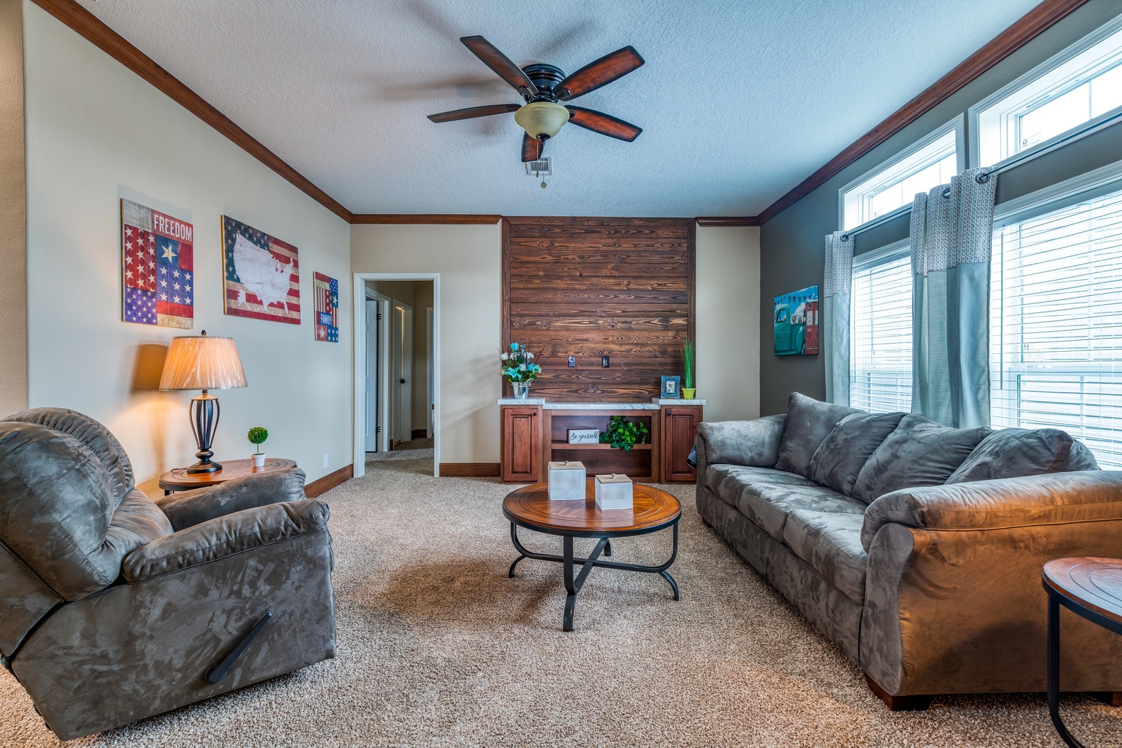Cozy living room with gray couches, brown armchair, round wooden coffee table, and patriotic wall art. Large windows and a ceiling fan add brightness.