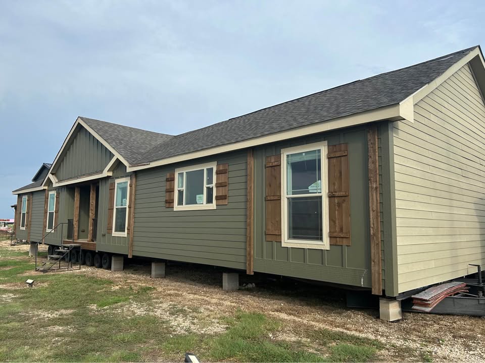 Row of modern modular ranch-style homes under construction featuring sage green vertical siding with beige trim accents, wooden brown shutters on large double-hung windows, gable roofs with dark asphalt shingles, covered front porches with wooden railings and steps, raised foundation on concrete block piers, light beige gable ends, spacious single-story manufactured bungalows exterior view in open construction site under partly cloudy sky