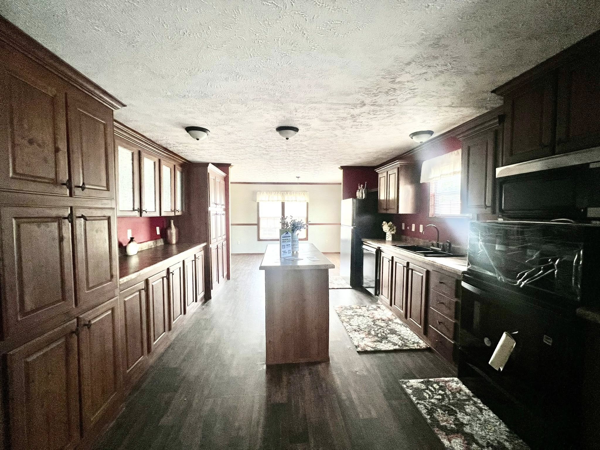 SEO-optimized alt text for a spacious manufactured home kitchen featuring extensive dark wood cabinetry, a central kitchen island, and a galley-style layout with modern black appliances; the high-ranking interior design showcases a functional and elegant floor plan with durable wood-look flooring, red accent walls, and recessed lighting that opens into a bright dining area, perfectly highlighting premium mobile home kitchen renovations and sophisticated modular home decor for maximum search engine visibility and ranking.

Would you like me to create SEO-optimized descriptions for any of the other rooms in this home?