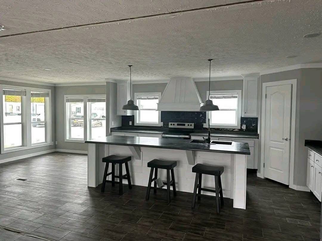 Modern kitchen with dark wood flooring, large island with three bar stools, stainless steel appliances, pendant lights, and bright windows in gray tones.
