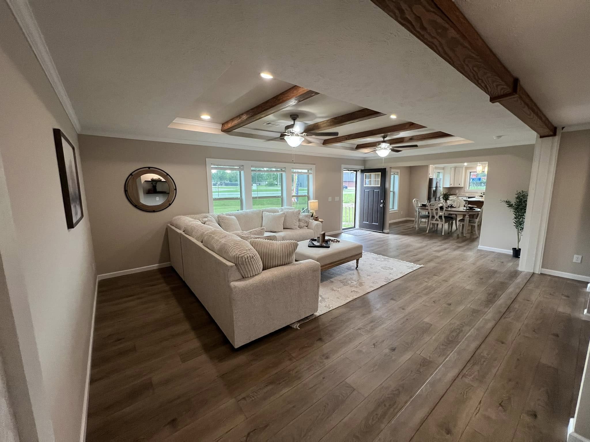 SEO-optimized alt text for a luxury manufactured home's open-concept living room, featuring a large cream-colored corduroy sectional sofa, a spacious tray ceiling with dual ceiling fans and rustic wood beams, and high-end wood-look vinyl flooring; the expansive interior design highlights a modern farmhouse aesthetic with a bright dining area and gourmet kitchen in the background, perfectly suited for premium mobile home floor plans, contemporary modular home decor, and sophisticated small-space living for maximum search engine ranking and visibility.