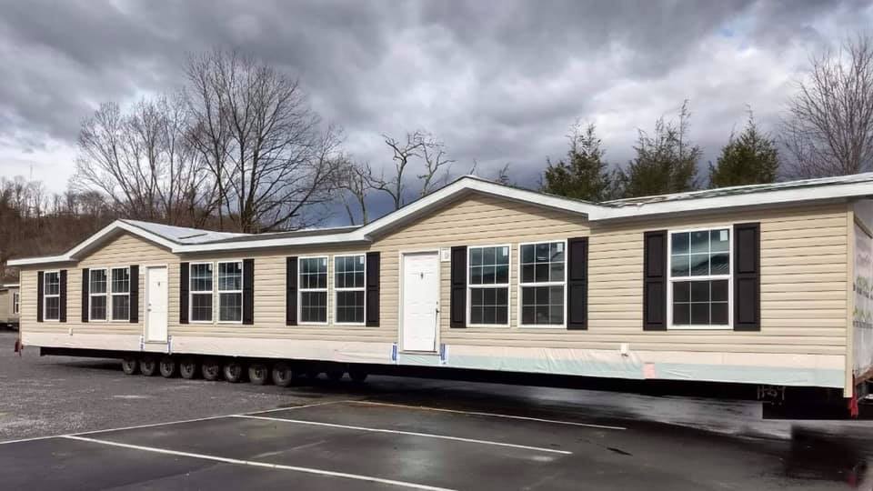 Modern double-wide manufactured home being transported on a multi-axle trailer, beige vinyl siding with black shutters, white trim, gable roof design, newly built modular home ready for delivery and setup, 2025 model mobile home in transit under cloudy sky