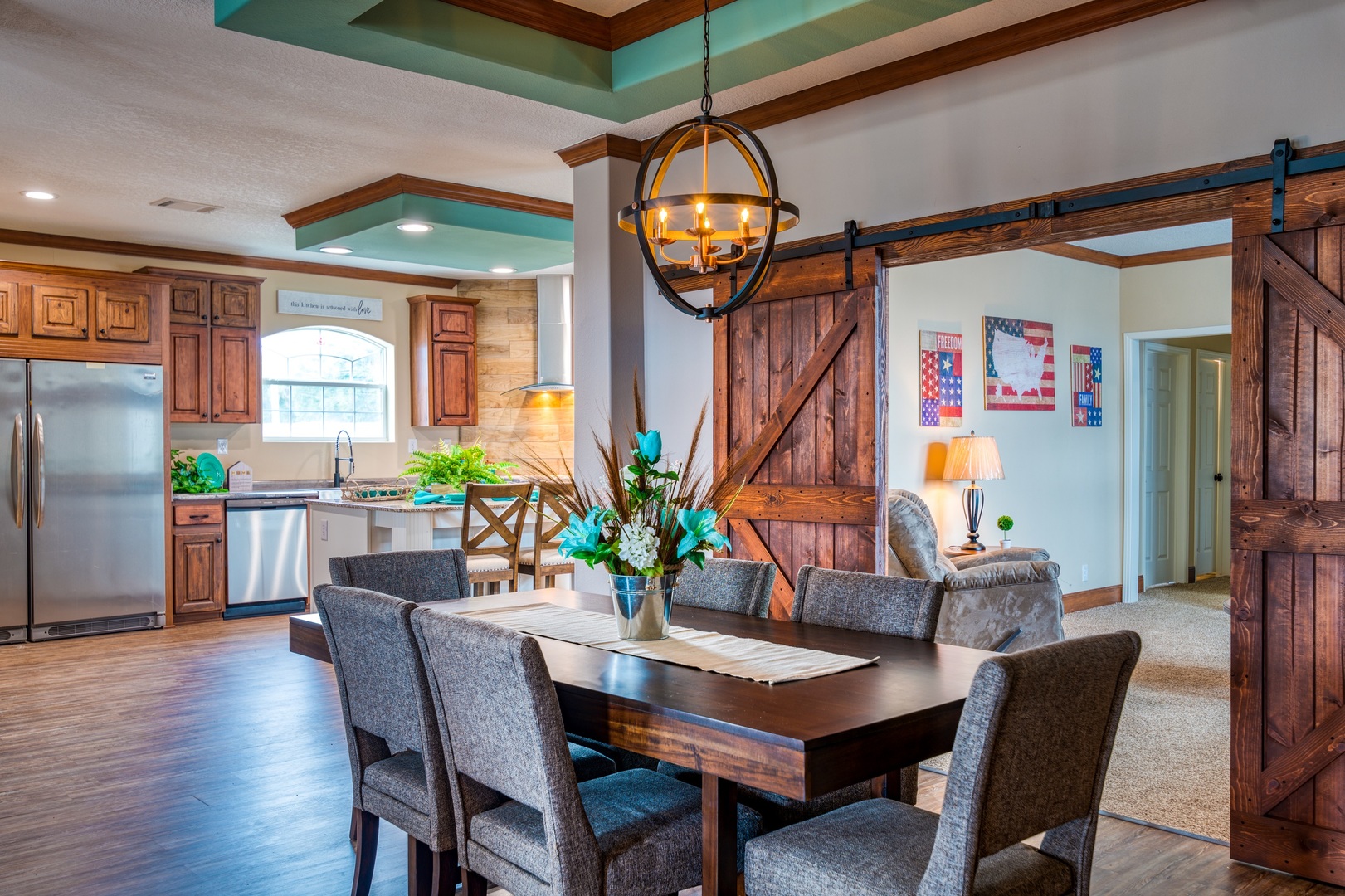 Open-concept dining and kitchen area featuring a dark wood dining table with upholstered chairs, a striking globe chandelier, rich wood cabinetry, stainless steel appliances, granite countertops, a rustic barn door, and patriotic wall decor, all under a uniquely accented ceiling.