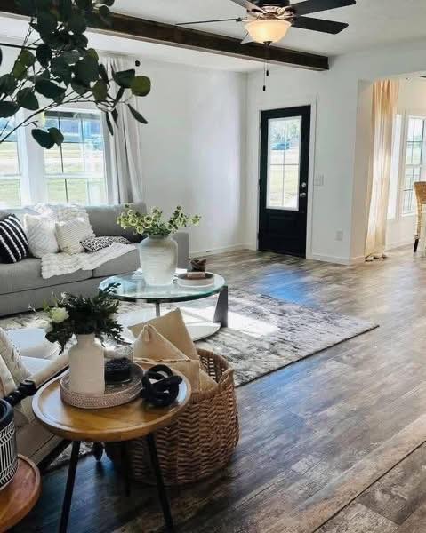 Inviting open-concept living room in a modern farmhouse modular home featuring bright white walls and vaulted ceilings with exposed dark wooden beams, elegant black ceiling fan with integrated light, plush light gray sectional sofa adorned with cozy white throw pillows and textured blankets, large glass-top round coffee table on a neutral patterned area rug over rich hardwood-look flooring, abundant natural light pouring through sheer white curtains on oversized windows and French doors leading to outdoor views, potted eucalyptus greenery, fresh sunflowers in white vase, woven baskets with decor accents, black entry door with glass panels, seamless flow to adjacent dining and kitchen areas creating a warm spacious family-friendly gathering space perfect for relaxation and entertaining in this dream affordable manufactured home.