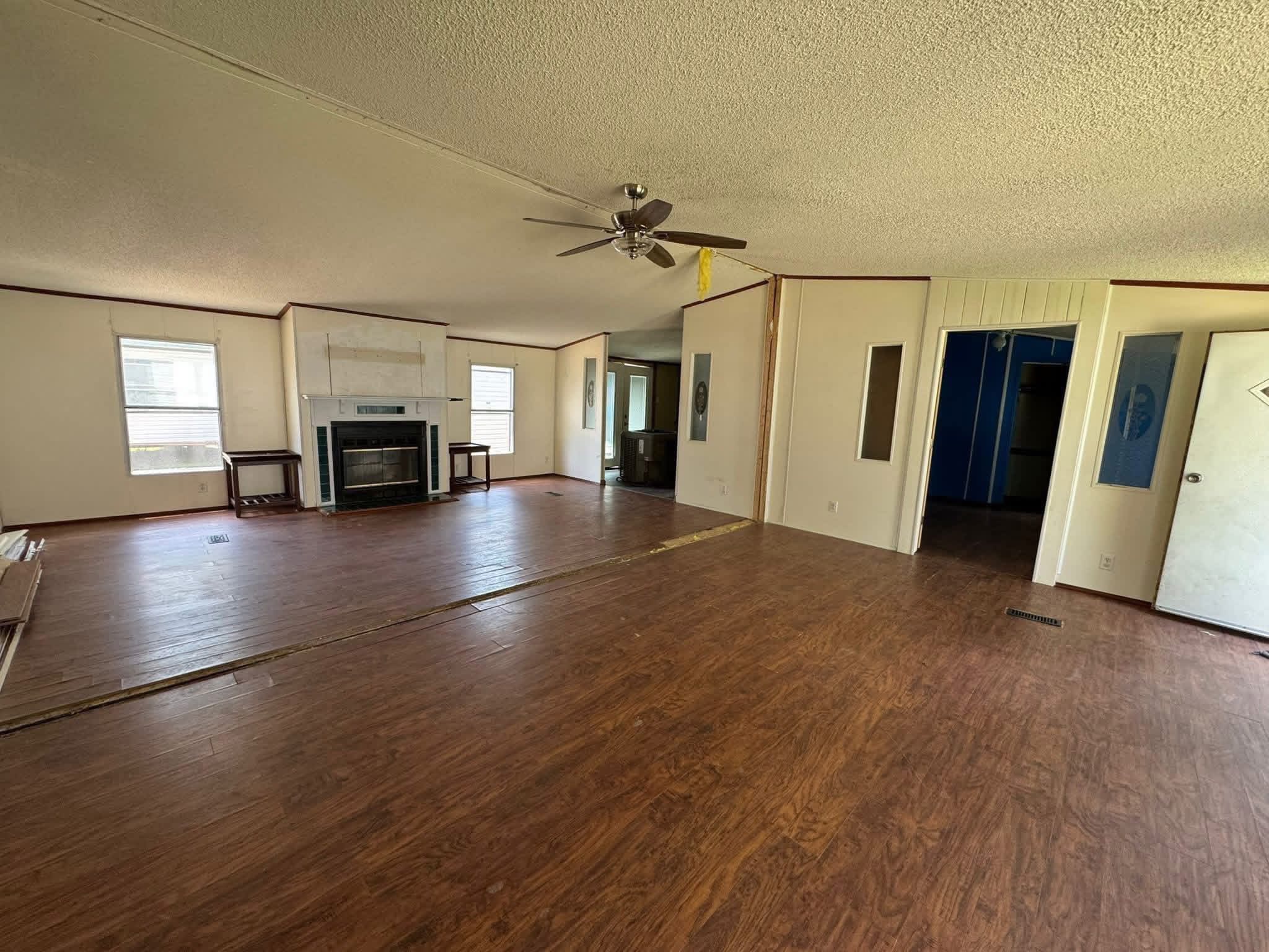 SEO-optimized alt text for an open-concept living room in a spacious manufactured home featuring wide-plank wood-look flooring and a modern white-mantel fireplace with a black insert; the high-ranking interior view showcases a versatile layout with neutral walls, a central ceiling fan, and multiple windows providing natural light, perfectly highlighting affordable mobile home floor plans, residential renovation ideas, and contemporary modular housing designs for maximum search engine visibility and ranking.