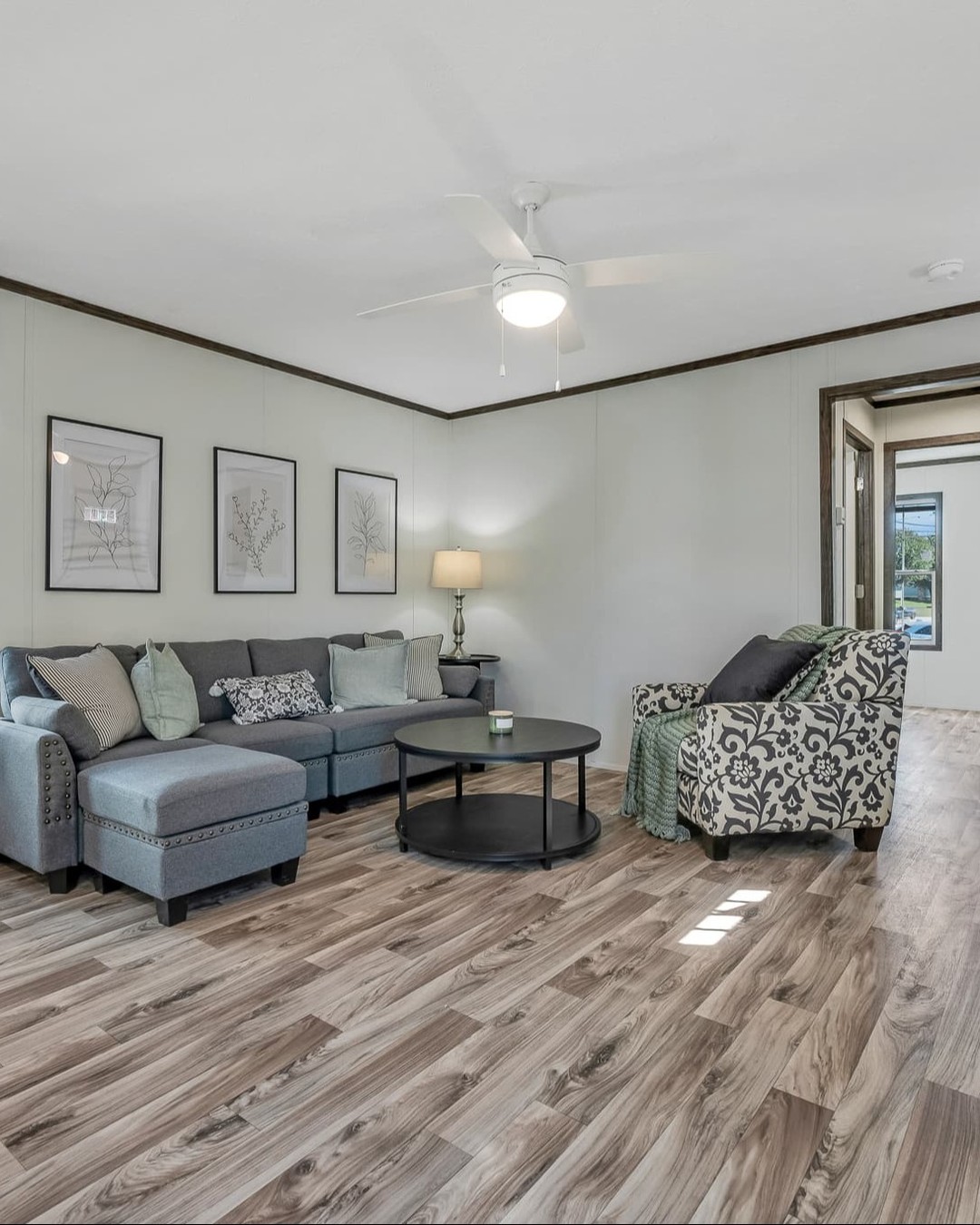 Cozy modern farmhouse living room in luxury tiny home or modular prefab cottage featuring spacious open layout with vaulted tray ceiling, elegant white ceiling fan with light kit, neutral light gray walls with dark brown chair rail molding, large sectional sofa in soft gray upholstery with nailhead trim and assorted throw pillows, matching patterned armchair with floral throw blanket, round black coffee table and side table, warm gray-brown luxury vinyl plank flooring, botanical wall art trio, bedside table lamp, bright natural light from adjacent windows, inviting comfortable seating area perfect for family relaxation in contemporary ranch-style manufactured home or vacation cottage interior design