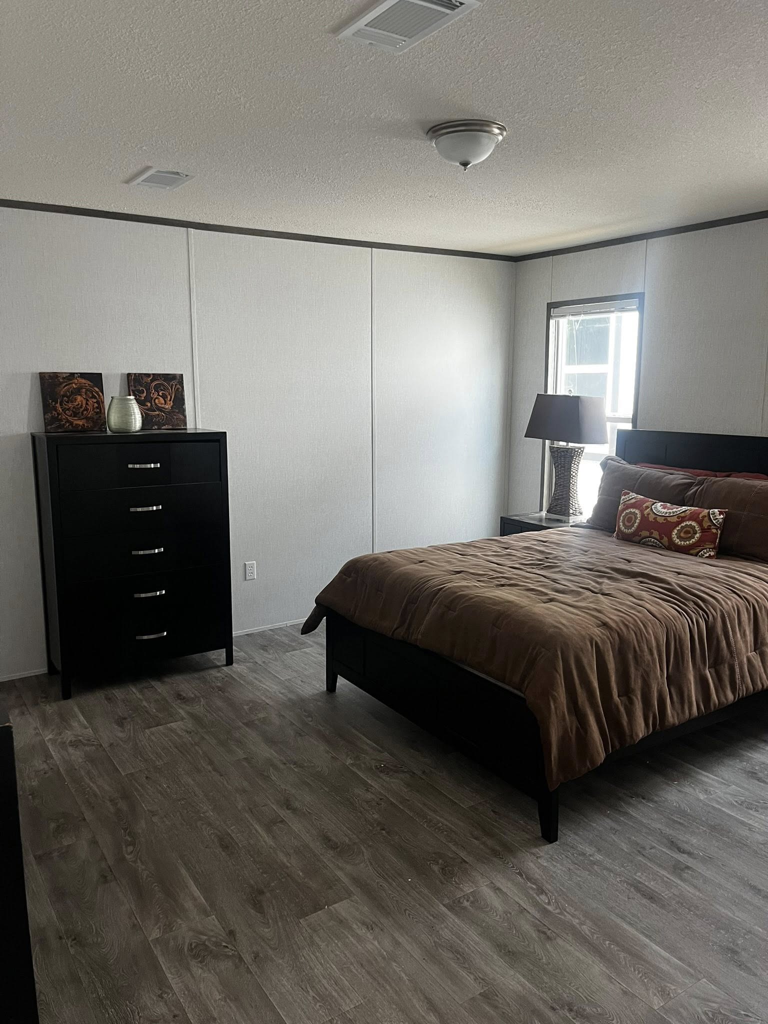 SEO-optimized alt text for a modern, high-ranking manufactured home bedroom featuring a dark wood queen-sized bed with a plush brown comforter and a matching tall black dresser with sleek silver hardware; the professional interior design showcases durable gray wood-plank flooring, light neutral walls with dark crown molding, and a large window providing natural light, perfectly highlighting luxury mobile home bedroom floor plans, contemporary modular home furniture ideas, and stylish residential interior finishes for maximum search engine visibility and ranking.