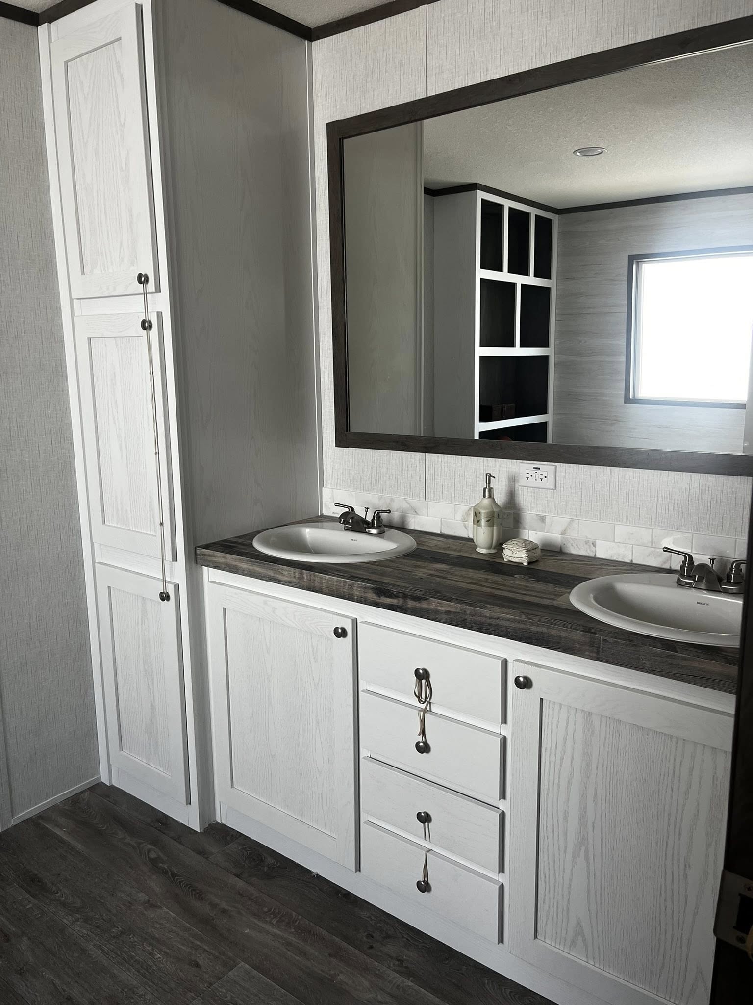 SEO-optimized alt text for a modern manufactured home bathroom featuring a double vanity with white shaker-style cabinets, dark wood-look countertops, and dual drop-in sinks set against a marble tile backsplash; the high-ranking interior design showcases a large dark-framed mirror reflecting built-in shelving, a tall white linen cabinet for storage, and durable gray wood-plank flooring, perfectly highlighting luxury mobile home bathroom floor plans, contemporary modular home cabinetry, and stylish residential master suite renovations for maximum search engine visibility and ranking.

Would you like me to describe the modern bedroom or the exterior of this home next?