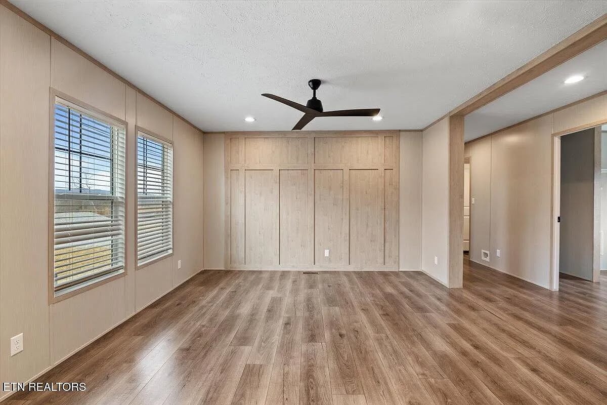 Spacious empty modern bedroom interior featuring large built-in wooden wardrobe closet, black ceiling fan, light wood-look vinyl plank flooring, beige paneled walls, large windows with blinds allowing natural light, recessed ceiling lights, and open doorway view – ideal for real estate listing in contemporary home design.