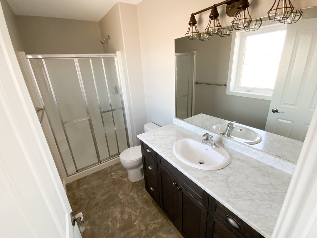 Modern luxury modular home bathroom interior featuring a sophisticated dark wood vanity with a high-end white marble countertop and integrated oval sink, accented by contemporary industrial cage-style vanity lighting, showcasing a pristine white toilet and a large glass-enclosed walk-in shower with neutral warm-toned walls and durable stone-look flooring for a 100% move-in ready turn-key residential design.