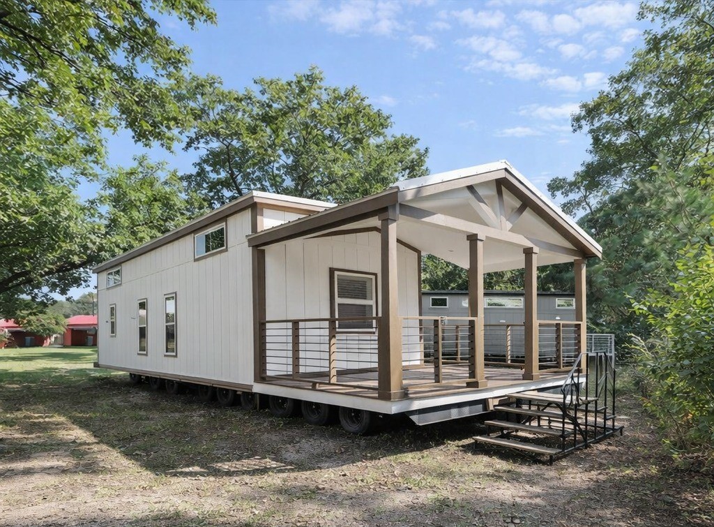 Modern luxury single-wide manufactured home for sale featuring a high-end white vertical siding exterior with a large covered front porch, exposed wooden beams, and a vaulted gable roof, set on a multi-axle chassis for a sophisticated move-in ready turn-key residential design perfect for a tiny home lifestyle or vacation property.