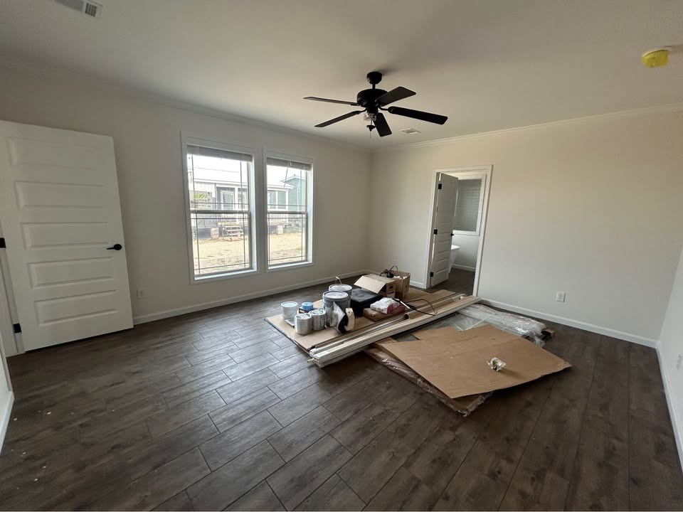 Spacious empty secondary bedroom interior in new modular home under construction featuring light neutral beige walls with white trim, dark gray wide-plank luxury vinyl flooring, large double-hung windows with white blinds allowing abundant natural light, black industrial ceiling fan with lights, textured vaulted ceiling, white six-panel interior doors, construction materials including paint cans drop cloths cardboard and tools scattered on floor, open doorway to adjacent bathroom, bright unfurnished manufactured ranch-style bedroom view ready for personalization