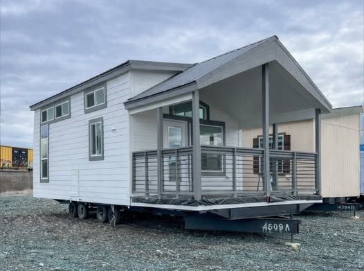 A modern tiny house on wheels with a white facade and large windows, featuring a covered porch with railings. It sits on a gravel lot under a cloudy sky.