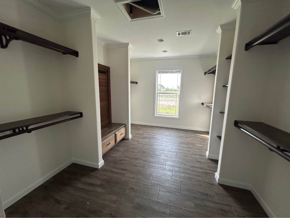 Spacious bright walk-in master closet interior in luxury modular home featuring extensive built-in dark walnut shelving and hanging rods with heavy-duty brackets on all three walls, large central island bench with under-storage drawers, neutral beige walls with crown molding and recessed lighting, dark gray wide-plank luxury vinyl plank flooring, single window providing natural daylight overlooking scenic outdoor view, vaulted ceiling with vent and can lights, elegant organized transitional farmhouse-style walk-in wardrobe closet space in new manufactured ranch home